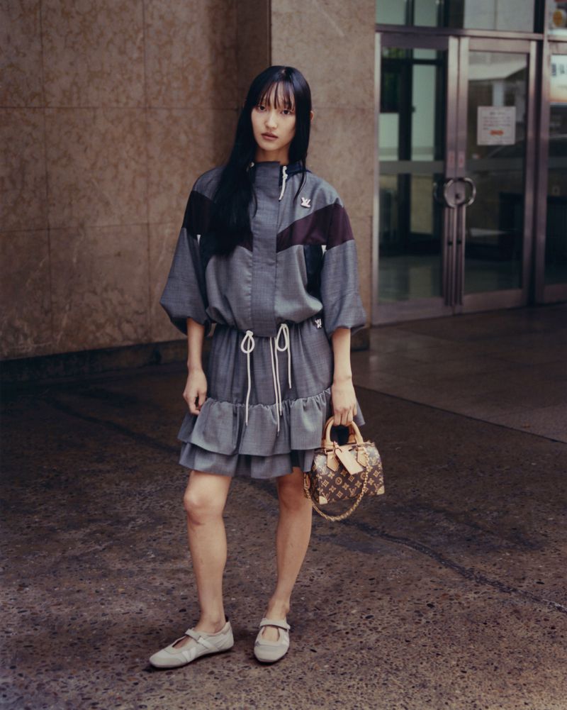 Model Carliane Paixao standing in front of a building exterior holding a monogram handbag and wearing a grey hooded dress