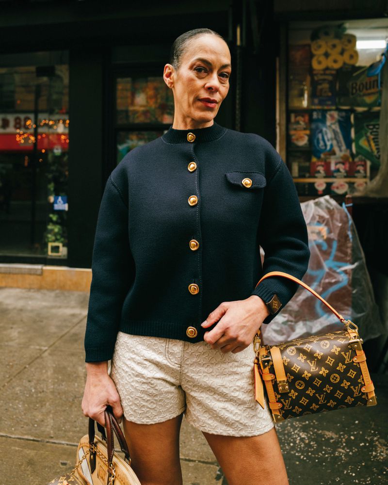 Model Carliane Paixao wearing a dark buttoned cardigan and light patterned shorts holding monogram handbags outdoors