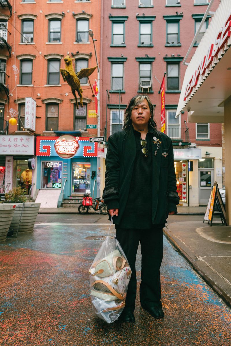 Model standing on a wet urban street holding a clear plastic bag filled with items, set against multi-story brick buildings.
