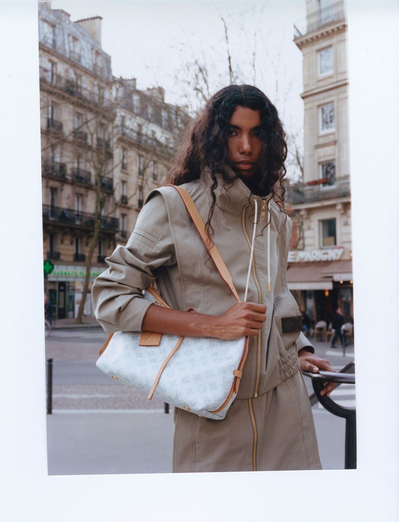 Woman with long dark hair wearing a tan jacket and skirt and carrying a purse stands on a sidewalk in front of buildings.