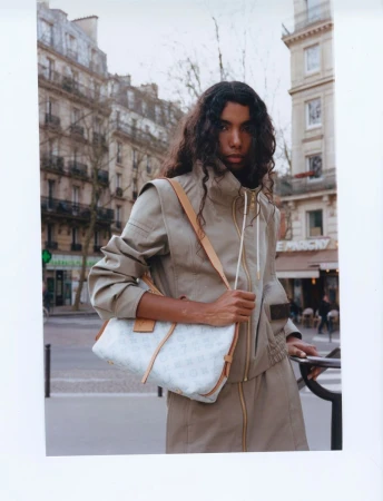 Woman with long dark hair wearing a tan jacket and skirt and carrying a purse stands on a sidewalk in front of buildings.