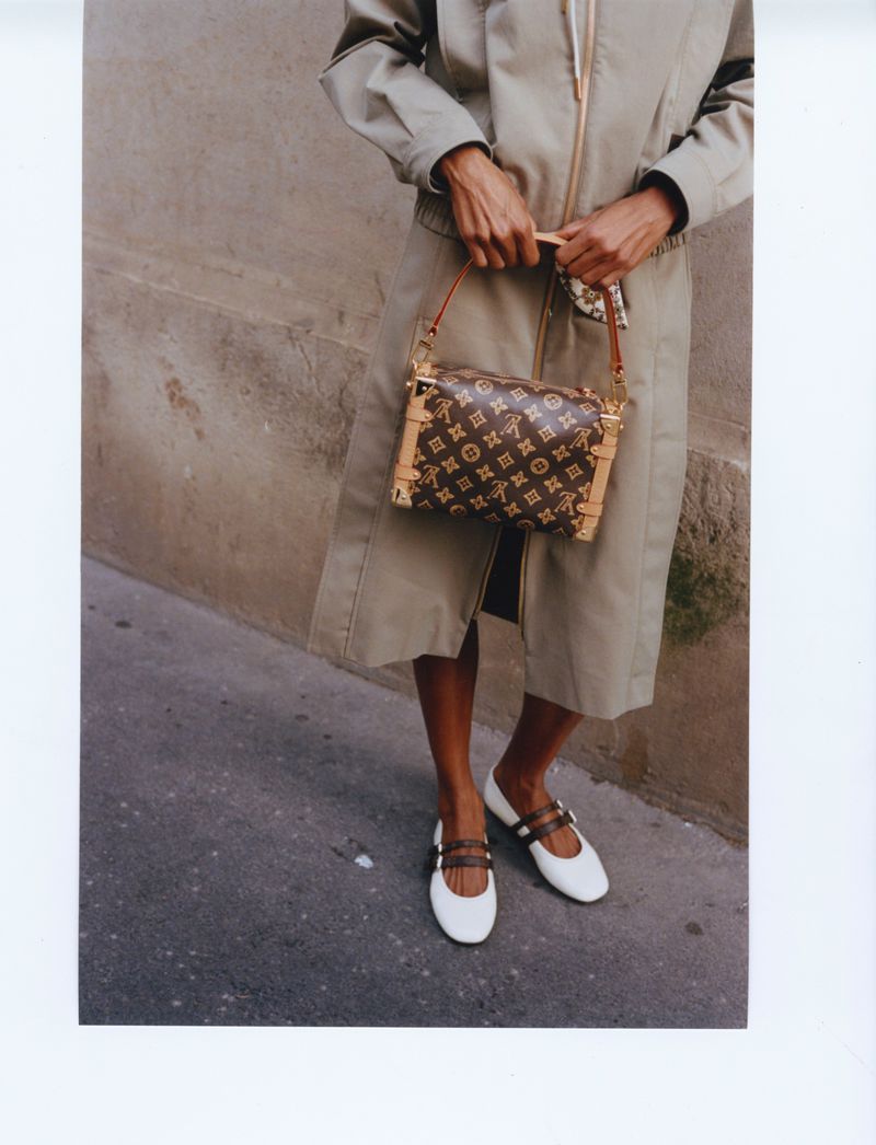 Model wearing a beige coat and white shoes holding a monogram handbag against an outdoor wall.