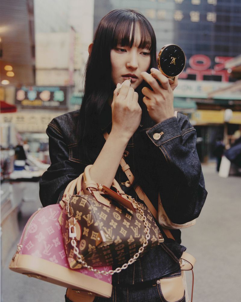 Model Carliane Paixao applying lipstick while holding luxury monogram handbags on a busy urban street