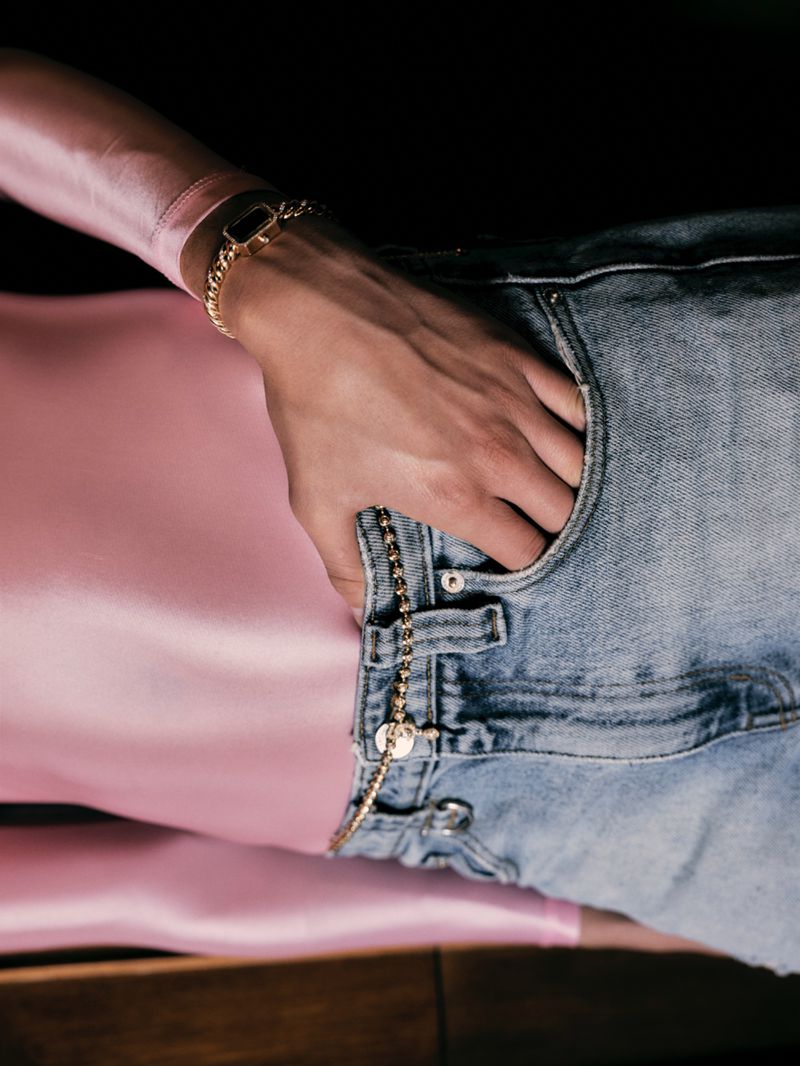 Close-up of a person wearing a pink long-sleeve top and blue denim jeans with a gold chain belt and wrist accessory.