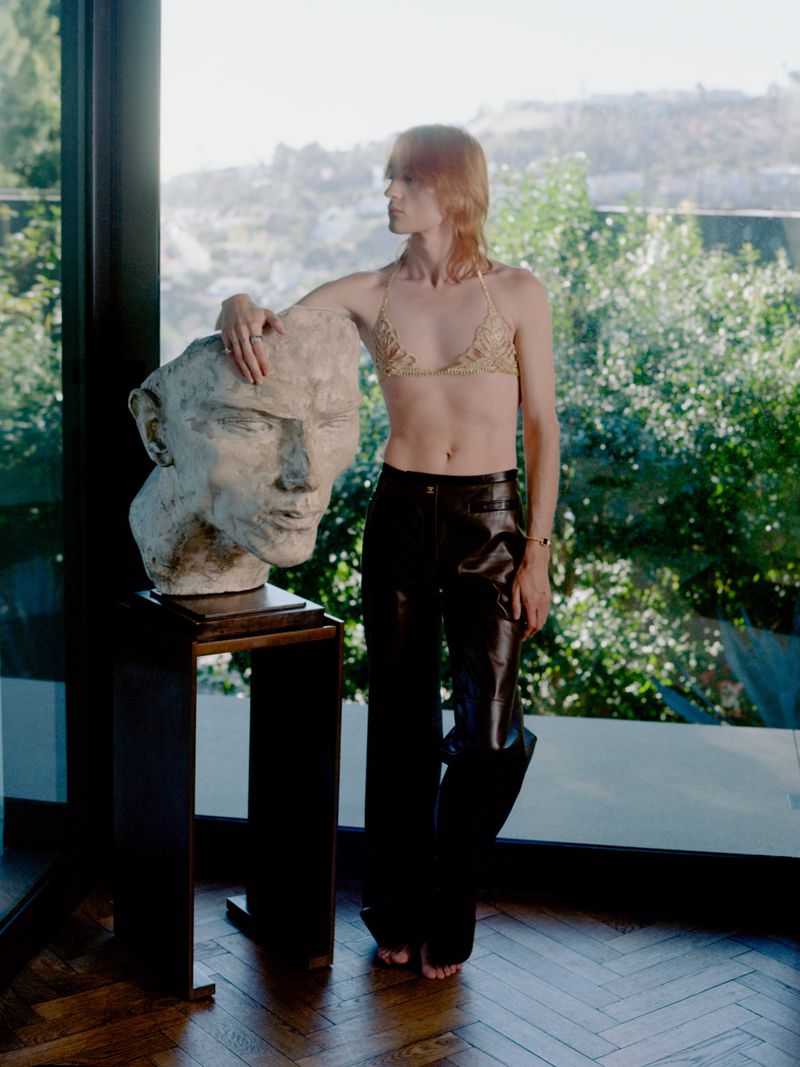 Model standing indoors beside a large sculpted head resting on a pedestal against a window backdrop with lush greenery