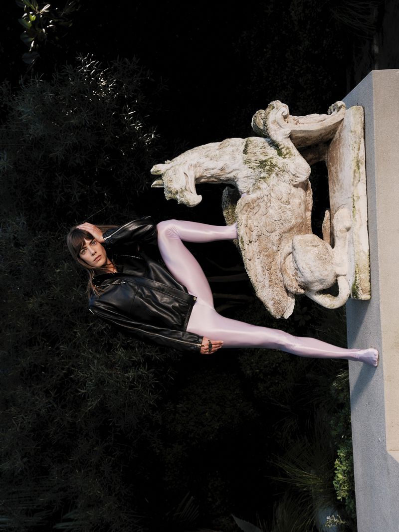 Model wearing a black leather jacket and shiny leggings posing with a stone sculpture outdoors for ODDA Magazine.