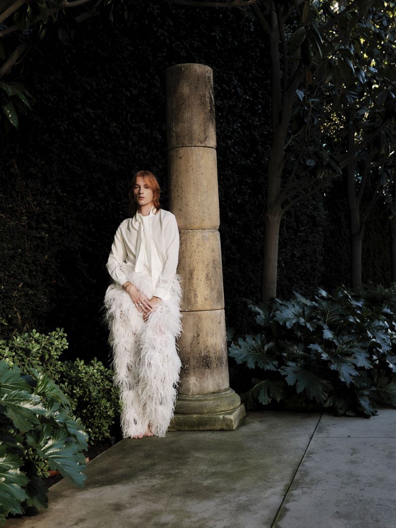 Model standing outdoors next to a stone column wearing a white blouse and feathery white trousers in a garden setting.