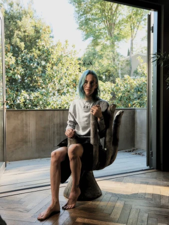 Woman with blue hair seated on a sculpted giant hand chair in front of an open balcony door