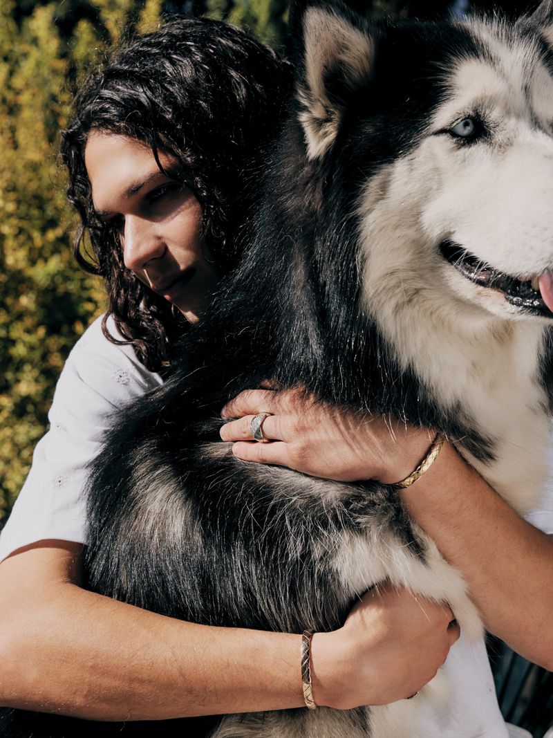 A man with long dark curly hair hugging a large black and white husky dog outdoors in an editorial portrait photograph.