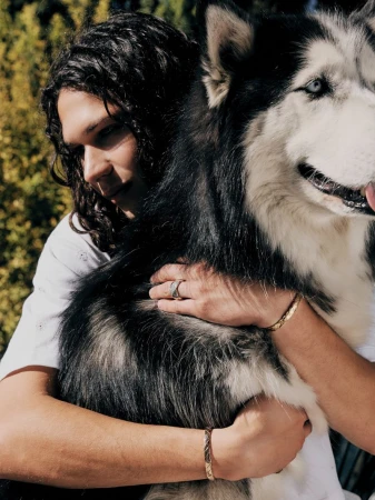 A man with long dark curly hair hugging a large black and white husky dog outdoors in an editorial portrait photograph.