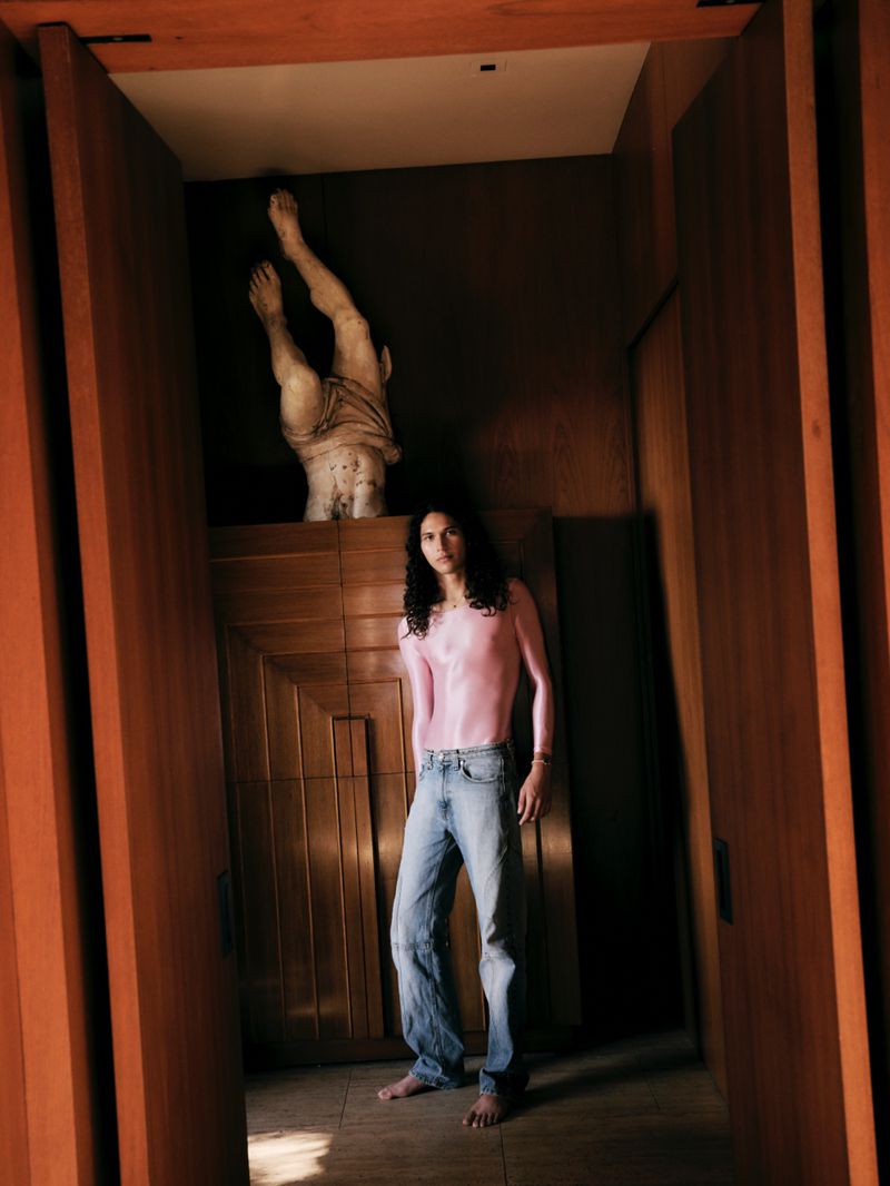 Model with long dark hair wearing a pink top and blue jeans standing in a wood-paneled room beneath a sculptural statue