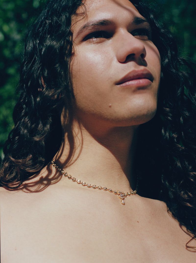 Close-up portrait of a person with long curly dark hair wearing a delicate gold pendant necklace against green foliage.