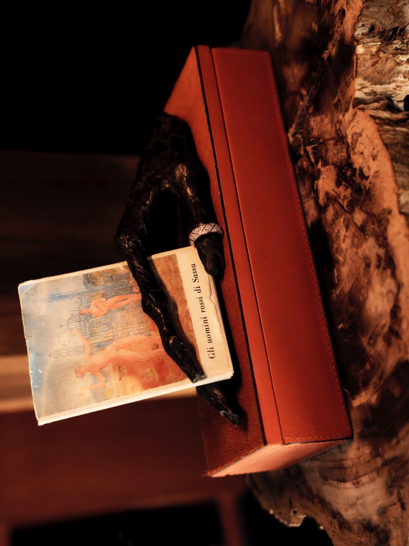 A sculptural black hand sculpture holding a small book against a brown leather box and wooden surface.