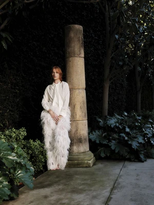 Model standing outdoors next to a stone column wearing a white blouse and feathery white trousers in a garden setting.