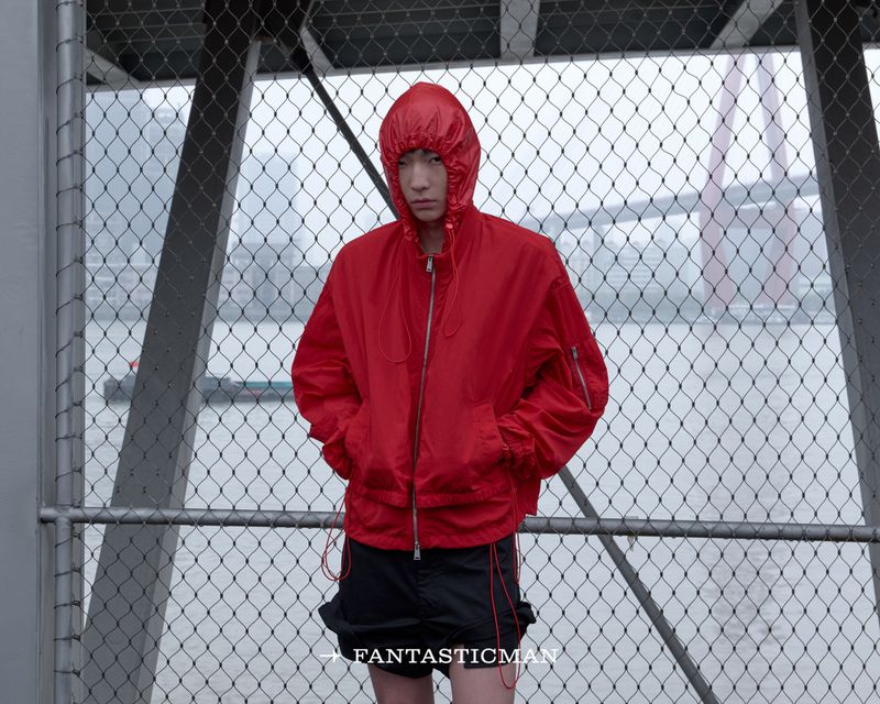 Model wearing a bright red hooded windbreaker and dark shorts posing behind a chain-link fence overlooking a river and bridge.