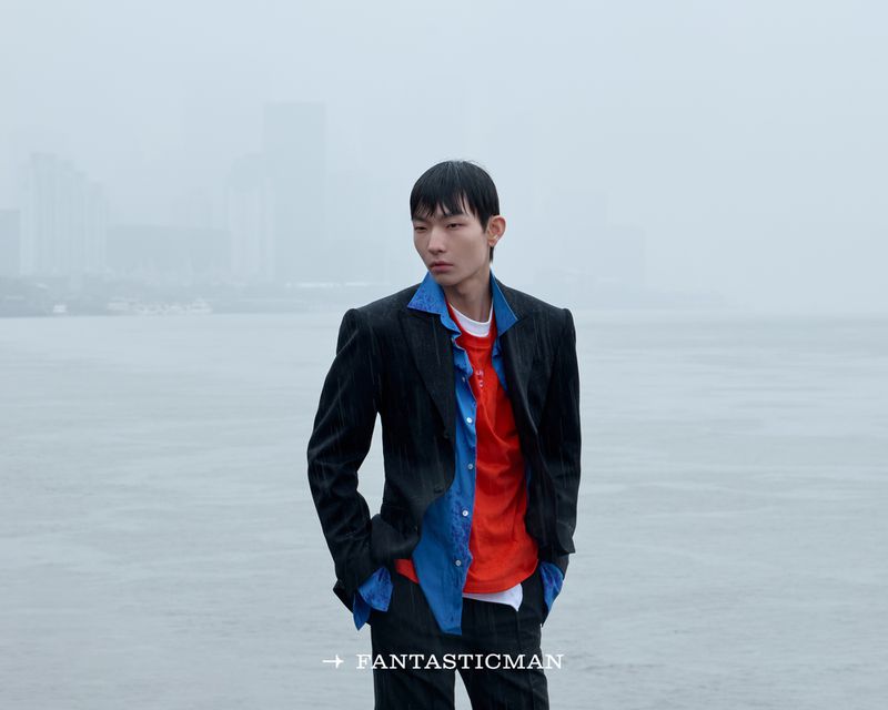 Asian male model in pinstripe blazer and layered blue shirt standing against an overcast waterfront backdrop.