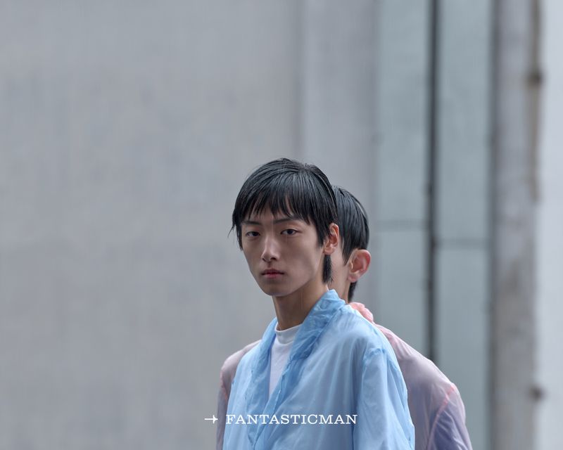 Two Asian men in sheer tops stand close together against a neutral grey background, showcasing a modern fashion aesthetic and minimal style.