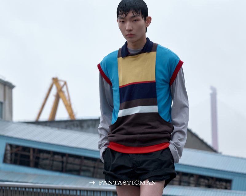 Model wearing a color-blocked vest over a long-sleeve top and dark shorts against an industrial background.