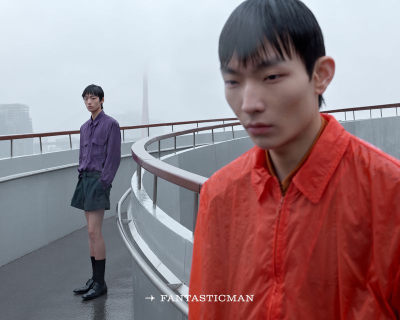 Two male models on a rooftop in the rain, one in a red jacket in the foreground and one in purple standing behind