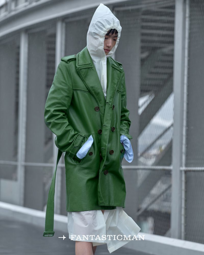 Model in a green belted coat and white hooded garment standing against an industrial bridge structure