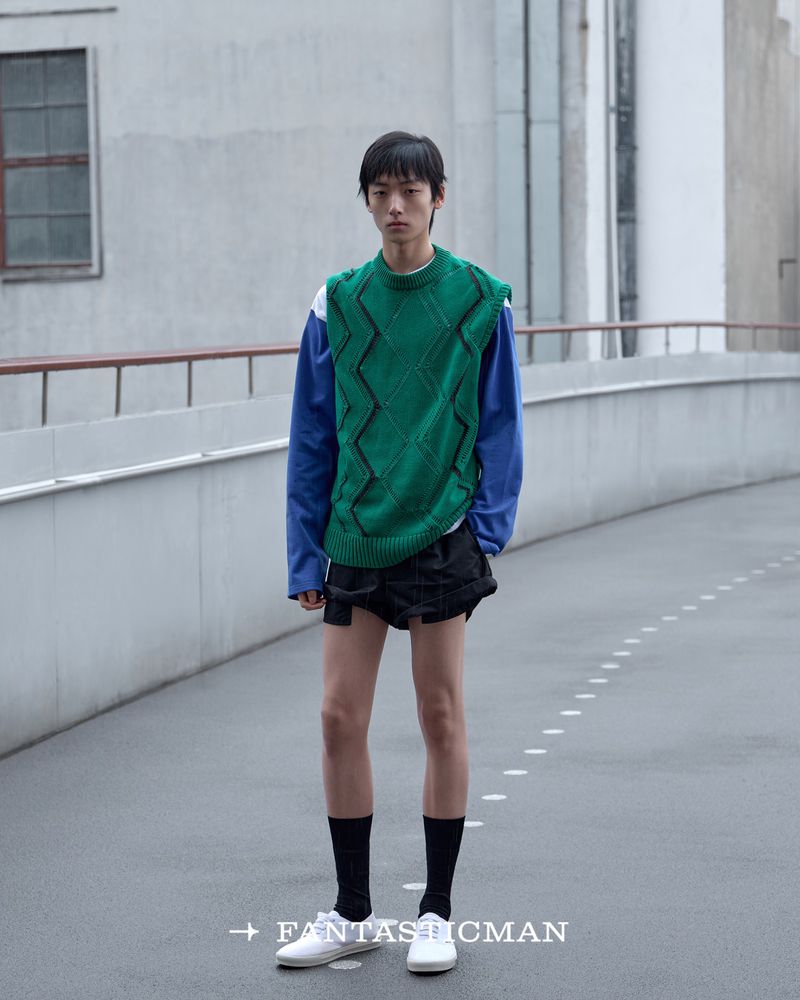 A young male model wearing a green sweater vest, blue long-sleeve shirt, and black shorts on an outdoor pathway.