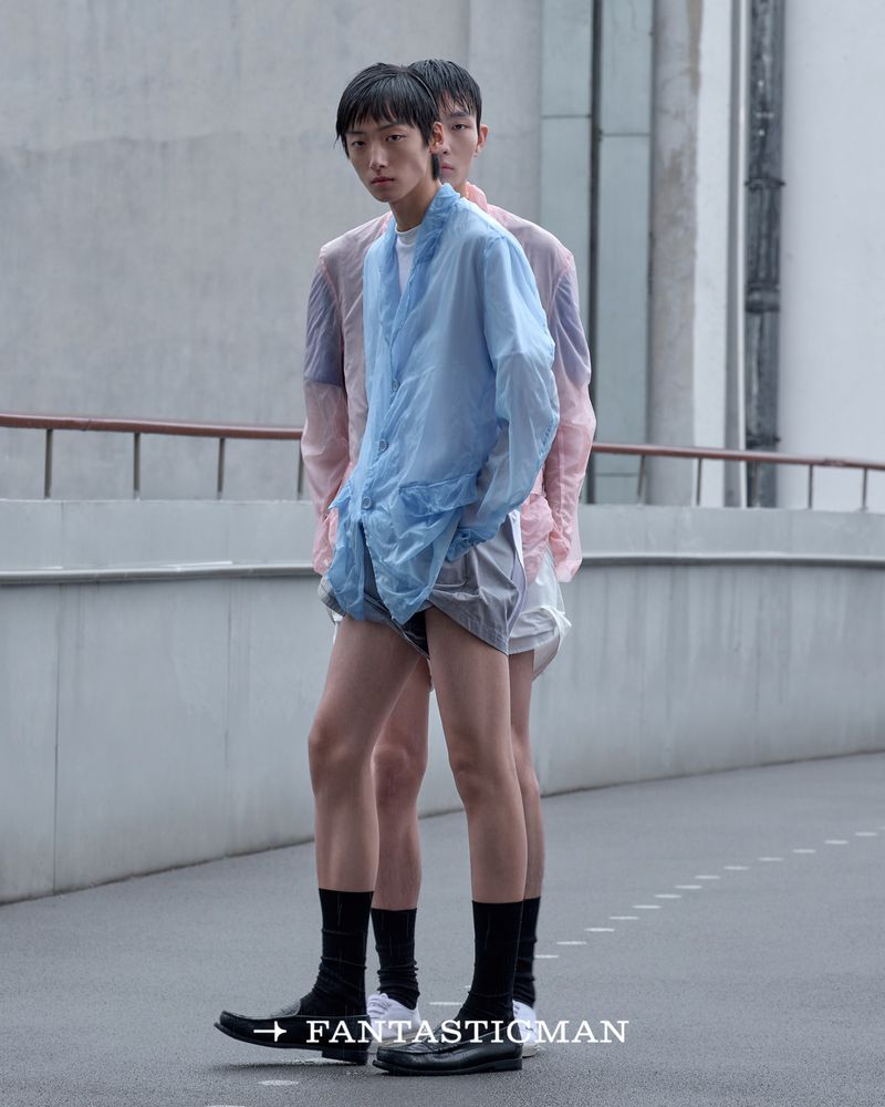 Two male models standing on an outdoor paved platform wearing light translucent shirts, shorts, black socks, and loafers.