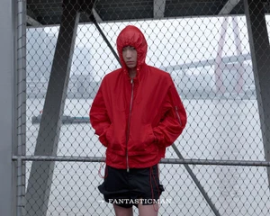 Model wearing a bright red hooded windbreaker and dark shorts posing behind a chain-link fence overlooking a river and bridge.