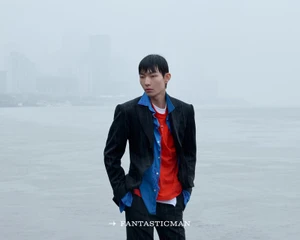Asian male model in pinstripe blazer and layered blue shirt standing against an overcast waterfront backdrop.