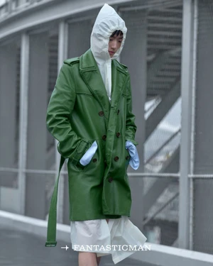 Model in a green belted coat and white hooded garment standing against an industrial bridge structure