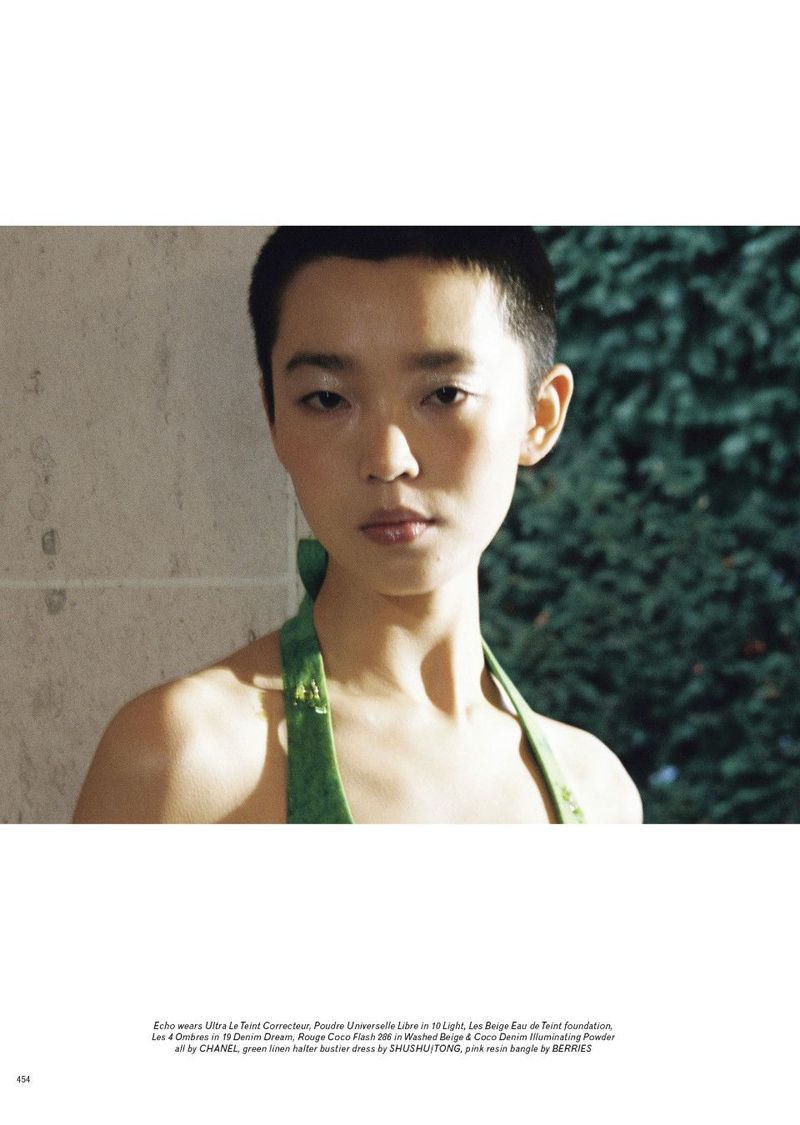 Close-up portrait of an Asian female model with a buzzcut, wearing a green halter top, posing in front of a leafy green background.