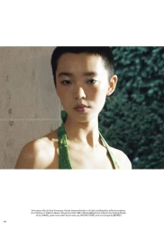 Close-up portrait of a model with short dark hair wearing a green halter dress against a stone wall and foliage background.