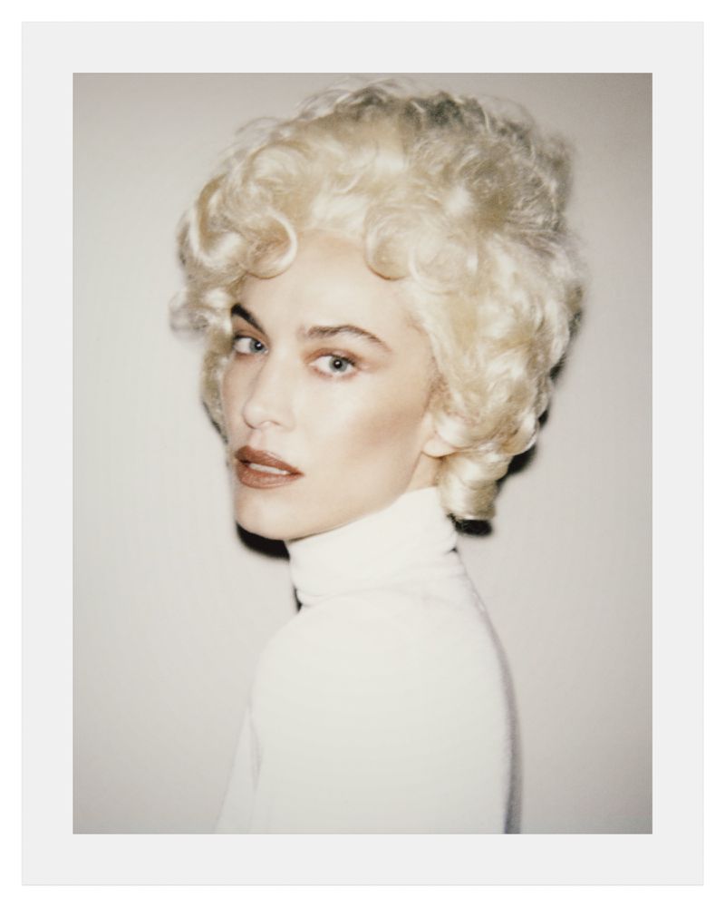 Portrait of Alexa Chung with voluminous blonde vintage hairstyle wearing a white turtleneck against a neutral backdrop