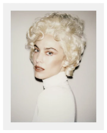 Portrait of Alexa Chung with voluminous blonde vintage hairstyle wearing a white turtleneck against a neutral backdrop