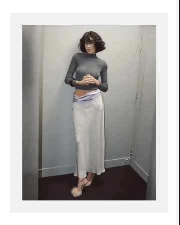 Alexa Chung wearing a grey turtleneck and long white skirt posing against an interior wall for Behind the Blinds