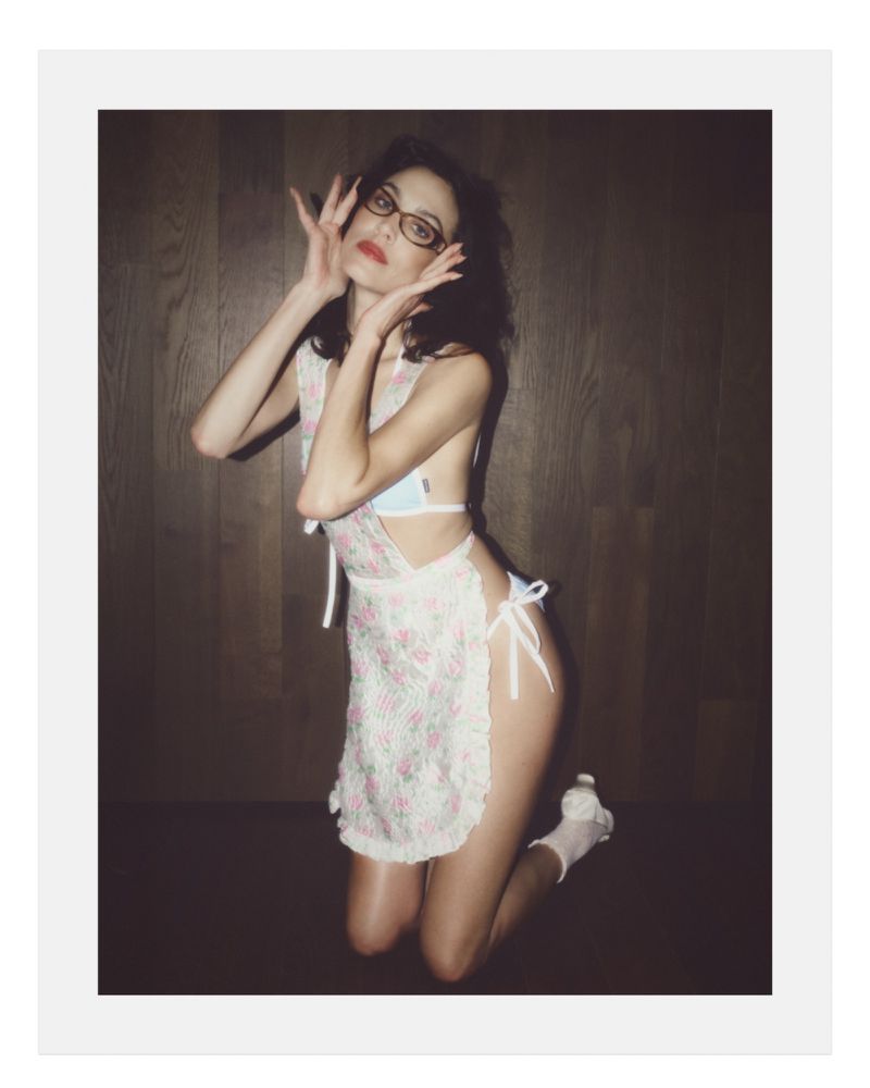 Alexa Chung wearing glasses and a floral apron while kneeling against a dark wood-paneled wall
