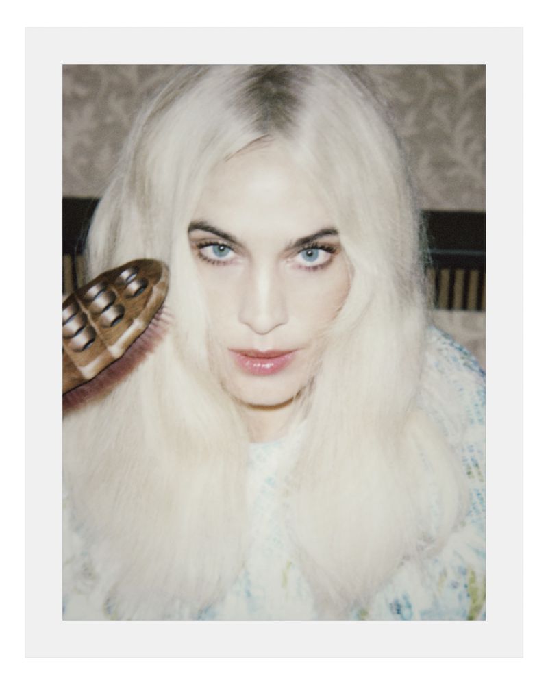 Blonde woman with blue eyes in a close-up headshot wearing a textured top with a floral wallpaper background and a hairbrush to the side.