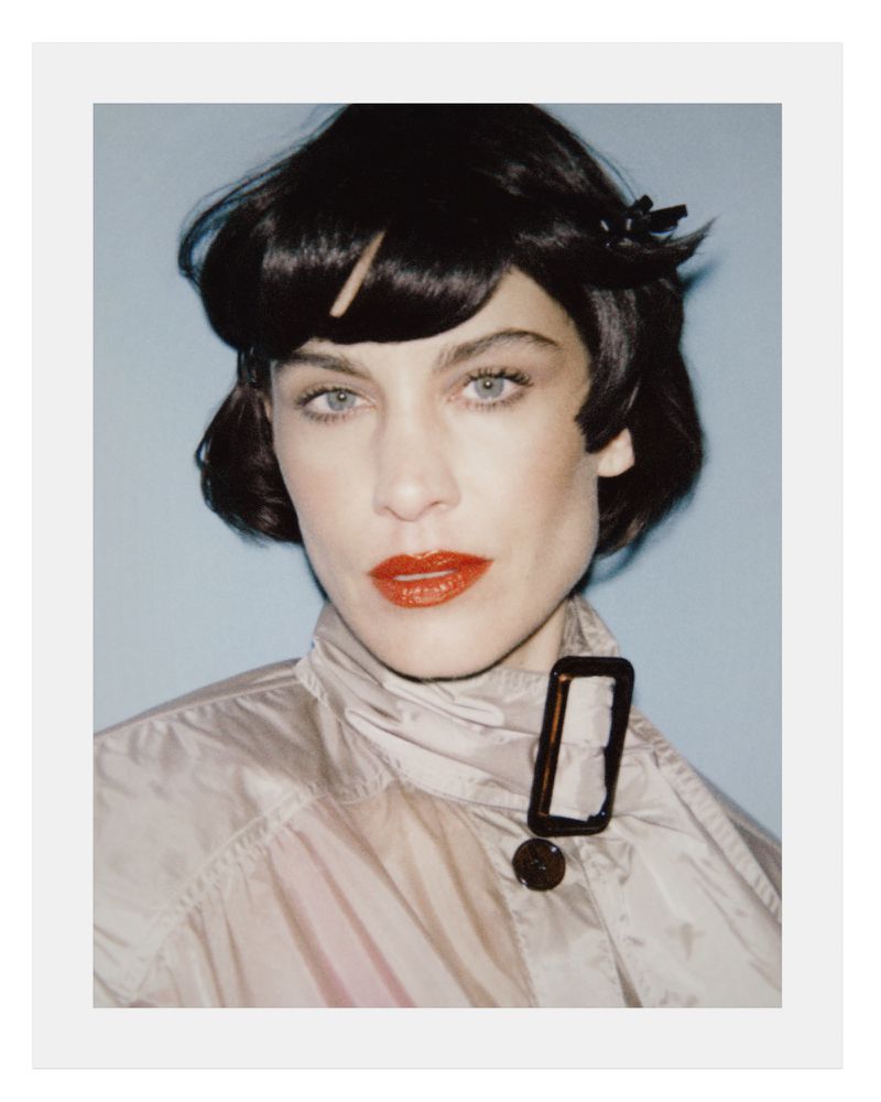 Portrait of Alexa Chung wearing a glossy beige trench coat with red lipstick and dark short hair against a grey background