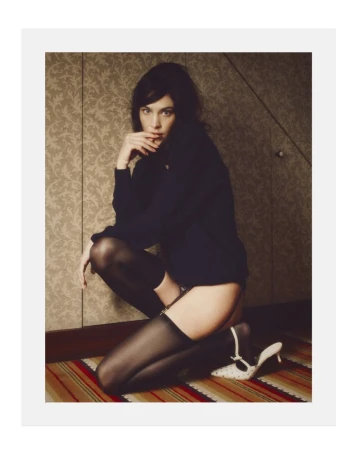 Alexa Chung kneeling indoors against a patterned wallpaper wall wearing dark clothing and stockings for Behind the Blinds editorial