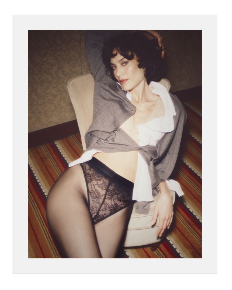 Alexa Chung seated in an upholstered chair wearing a grey cardigan, white collared shirt, and black lace briefs.