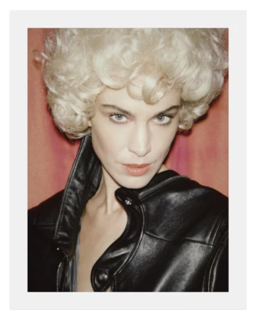 Alexa Chung wearing a black leather jacket and blonde curly wig against a reddish backdrop for Behind the Blinds.