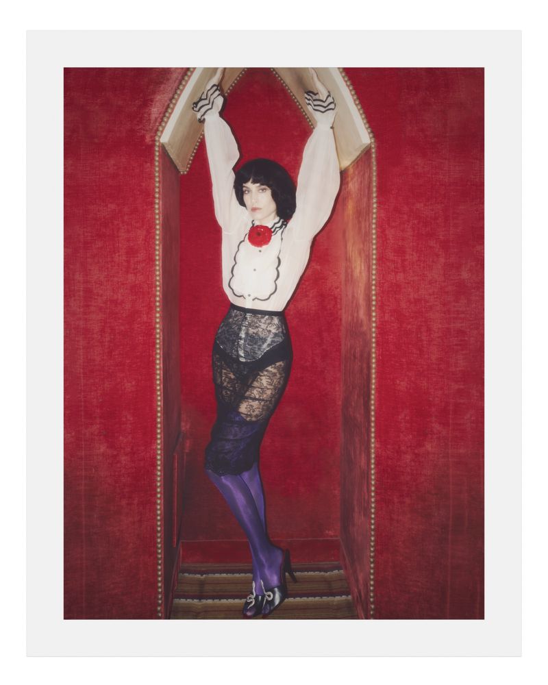 Alexa Chung standing with arms raised inside a red velvet alcove wearing a ruffled white blouse and black lace skirt