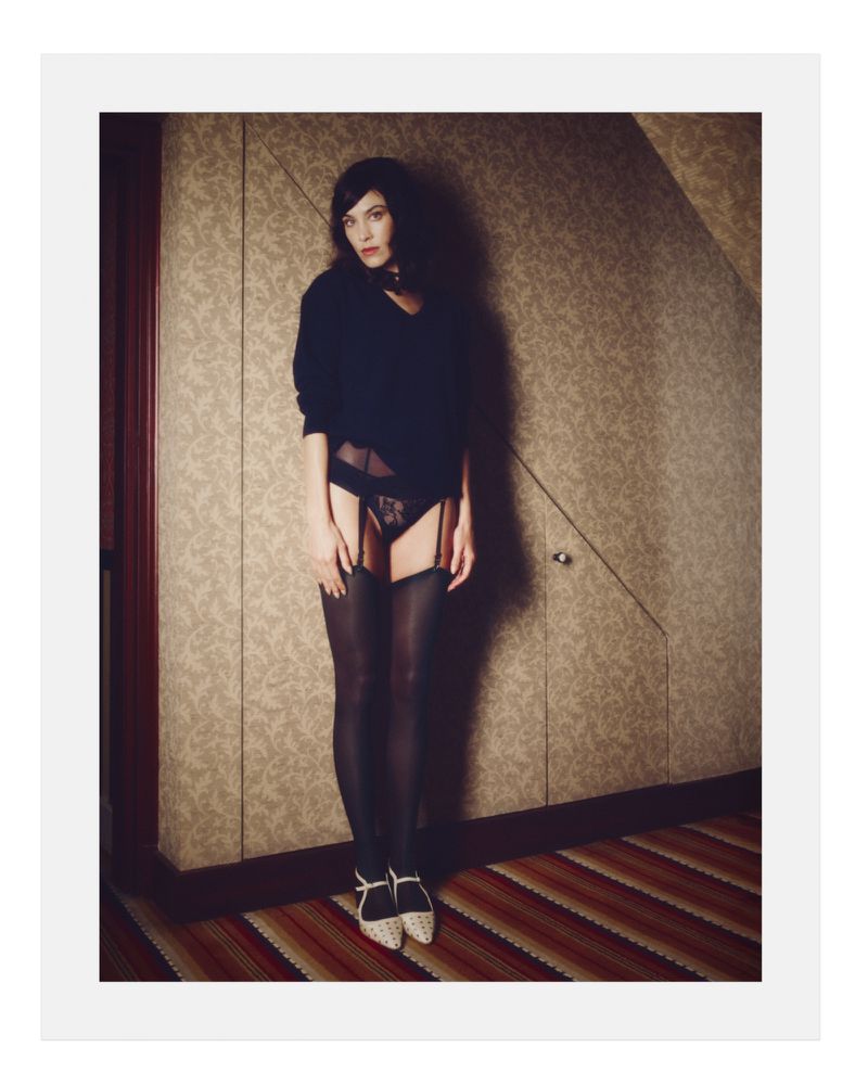 Alexa Chung posing in dark lingerie and sweater against patterned wallpaper for a Behind the Blinds editorial.