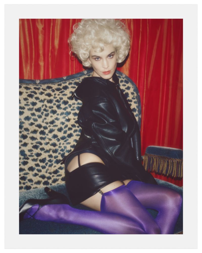 Alexa Chung wearing a blonde wig, black leather jacket, and purple stockings on a leopard-print sofa against red drapes.