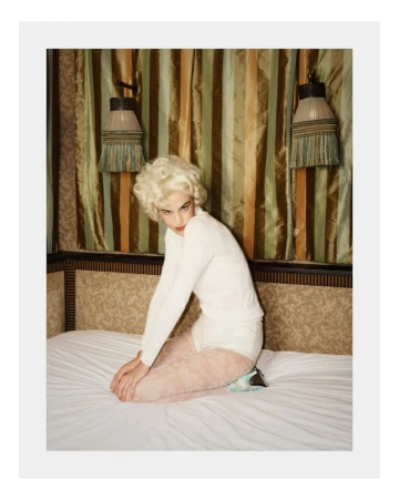 Alexa Chung kneeling on a white bed wearing a white long-sleeve top and patterned tights beneath green striped curtains