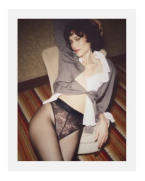 Alexa Chung seated in an upholstered chair wearing a grey cardigan, white collared shirt, and black lace briefs.