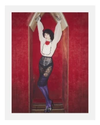 Alexa Chung standing with arms raised inside a red velvet alcove wearing a ruffled white blouse and black lace skirt
