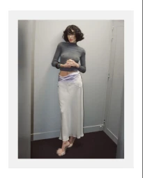 Alexa Chung wearing a grey turtleneck and long white skirt posing against an interior wall for Behind the Blinds