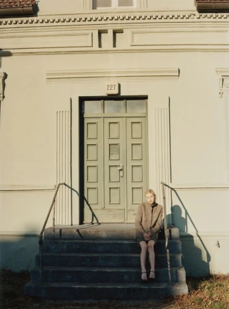 Young woman sitting on stone steps outside a residential building door, wearing a jacket and skirt