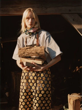 Blonde model holding a stack of cut logs in front of a rustic wooden structure, wearing a patterned skirt and light top.