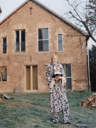 Model wearing a blue and white floral patterned coat and headscarf standing in front of a brick house for Achtung Magazine.